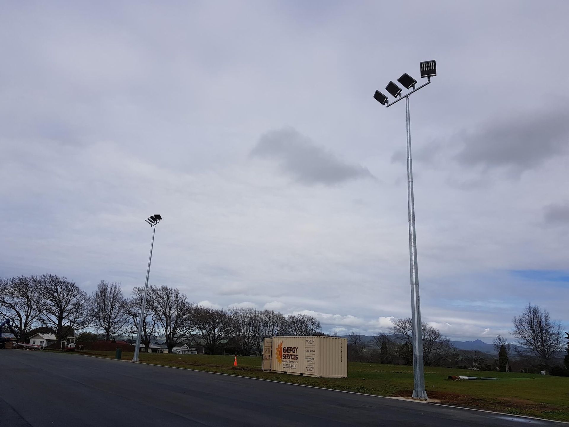 Two tall light poles with spotlights along a road, under a cloudy sky. A small wooden structure and trees are in the background.