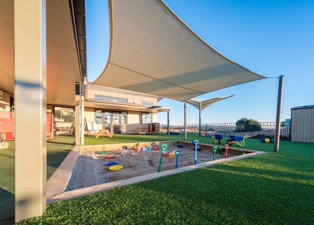 Outdoor play area at a childcare center with sandpit, artificial turf, and shade sails. Buildings are visible in the background.