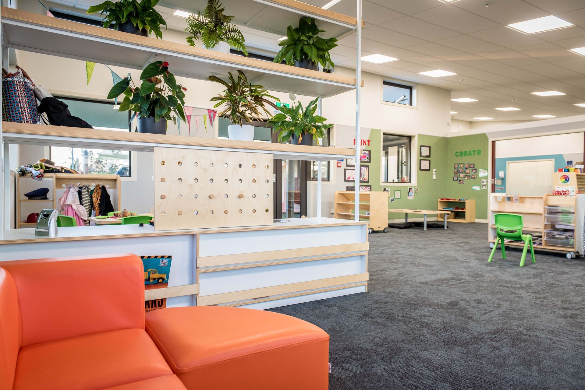 Interior of a brightly lit preschool classroom with orange seating, shelves holding plants, and various play areas.