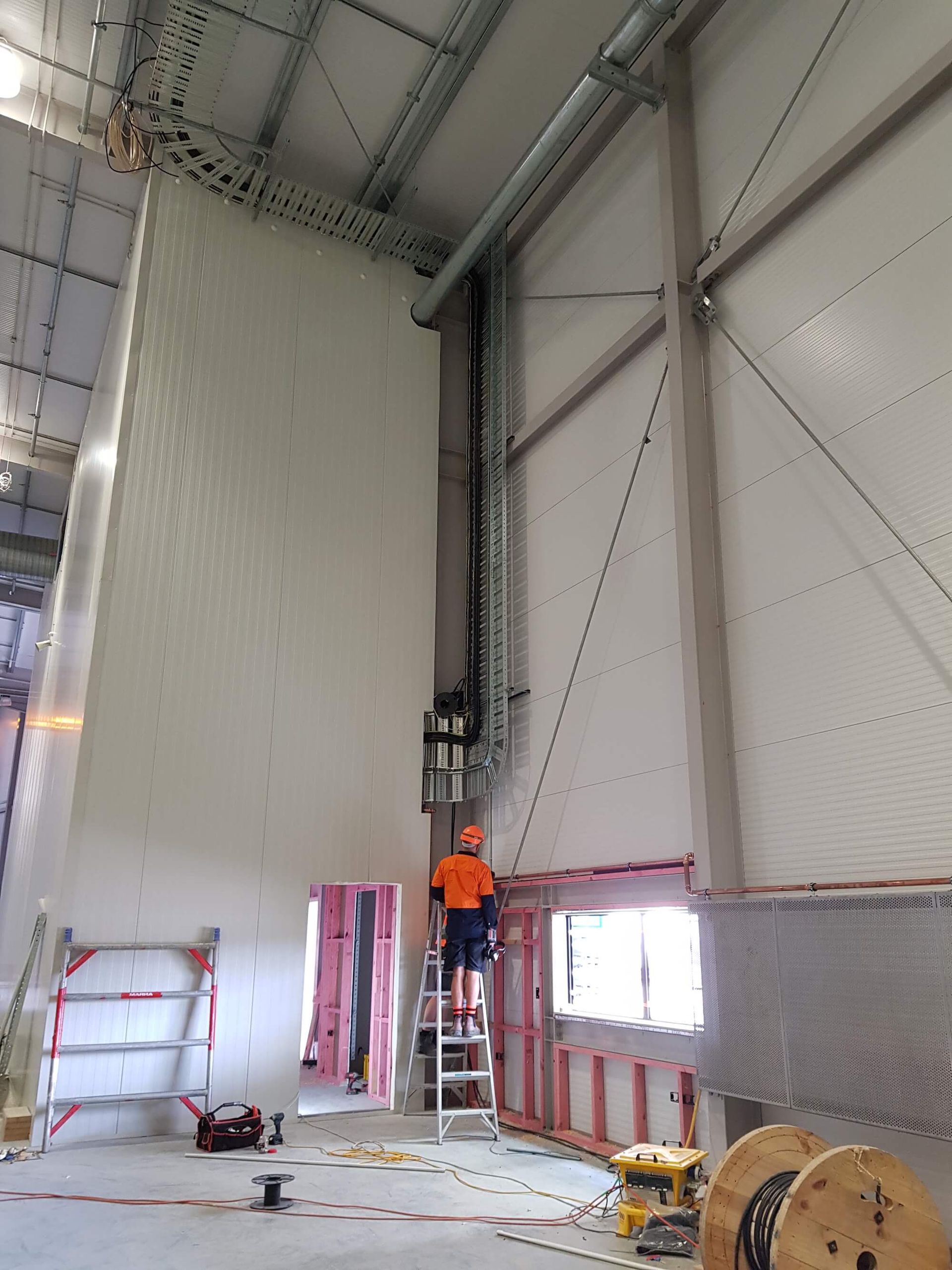 Construction worker on a ladder inside a large building, wiring near a high wall with insulated panels. Cables run up the wall to the ceiling.