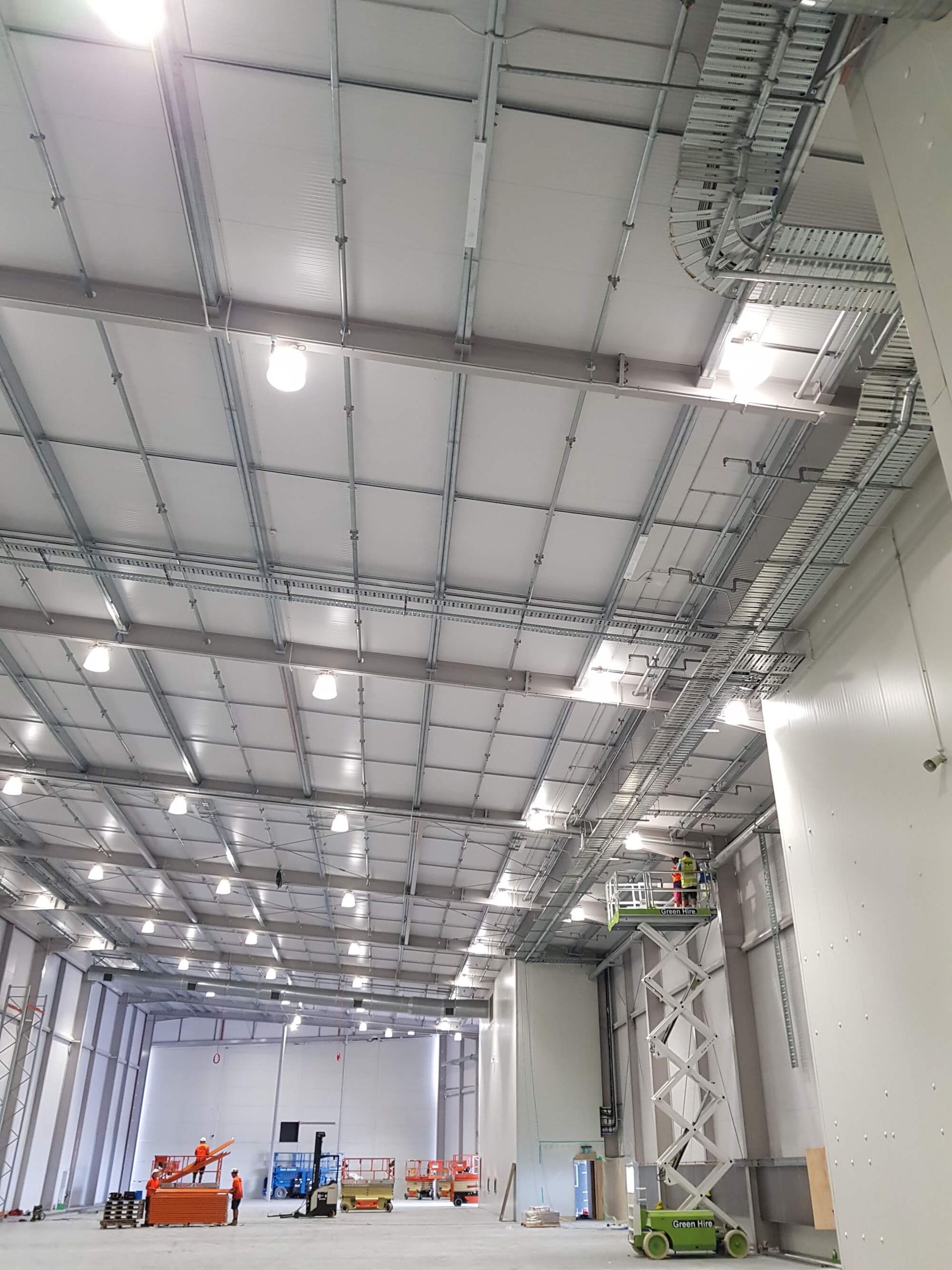 Interior view of a large, unfinished industrial space with metal framework ceiling and walls. Workers and equipment are present on the floor.
