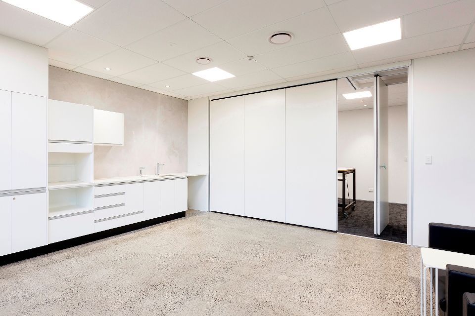 Bright, modern office space with white cabinetry, sliding partition doors, and a concrete floor. A doorway leads to another room with a table.