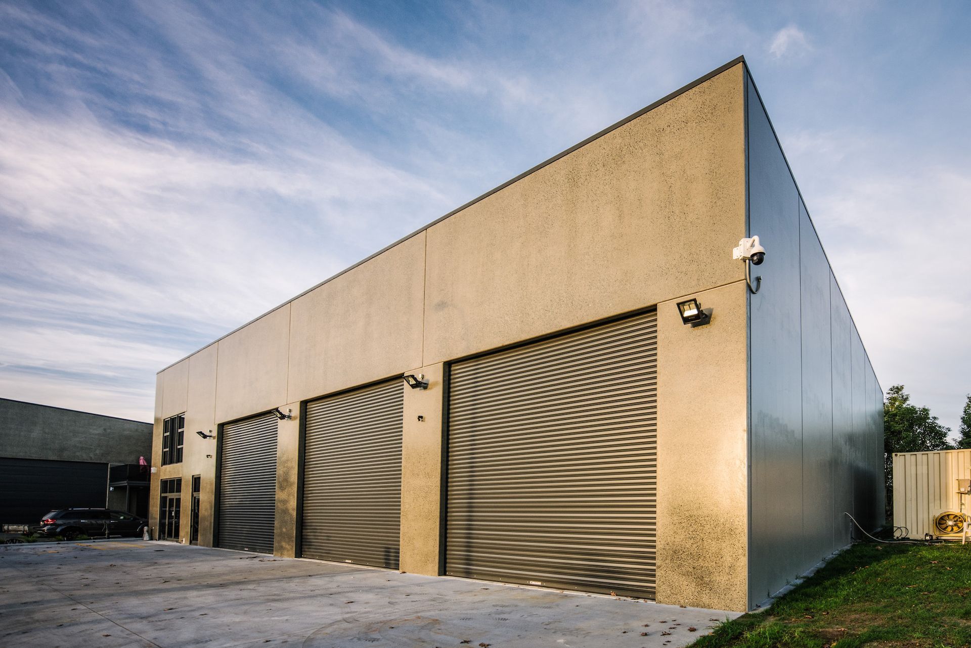 Exterior of a modern, tan-colored building with three large garage doors under a cloudy sky. Security cameras are mounted on the walls.