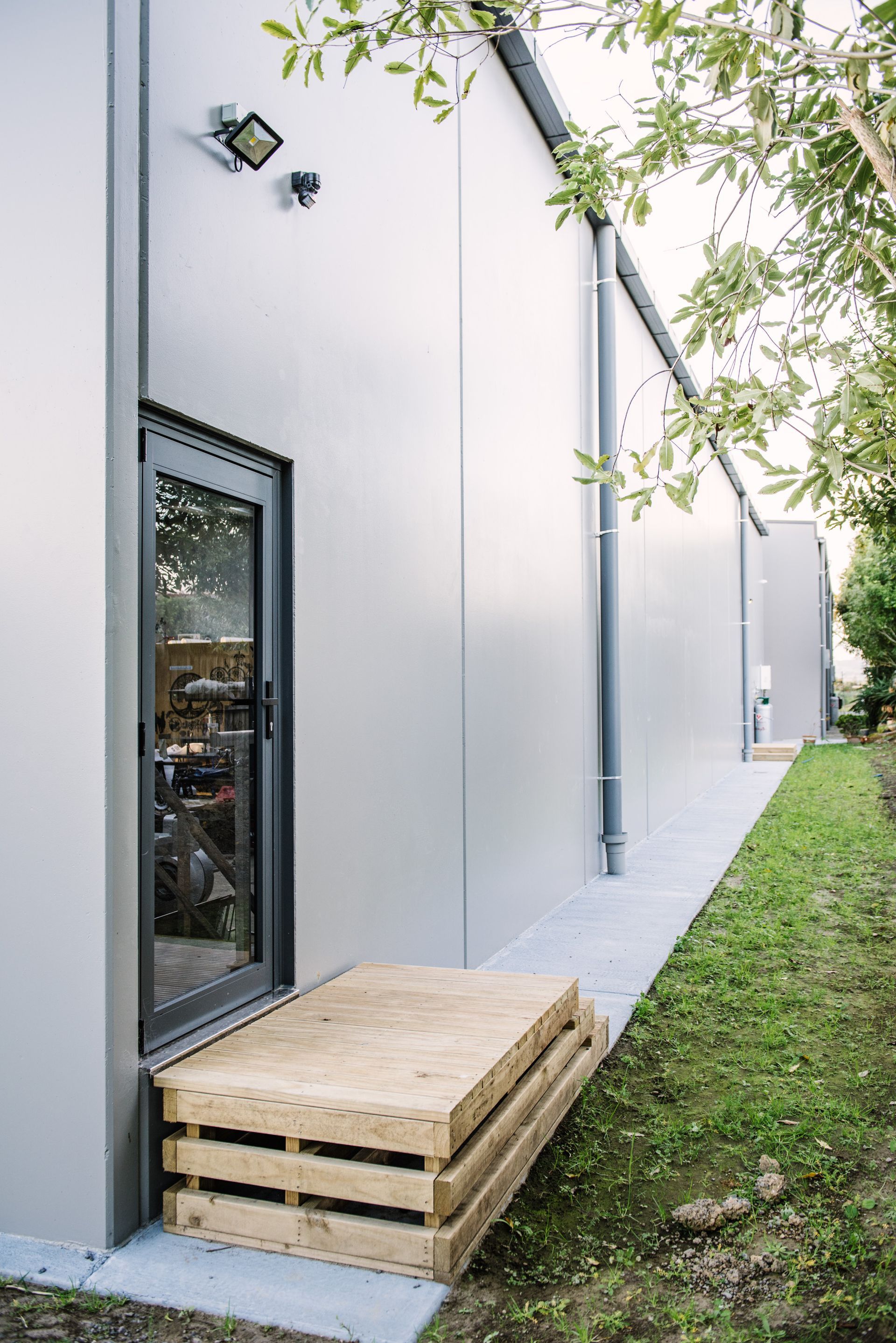 Exterior view of a building with a gray door and step, with a wooden pallet next to it. Green grass surrounds the building.