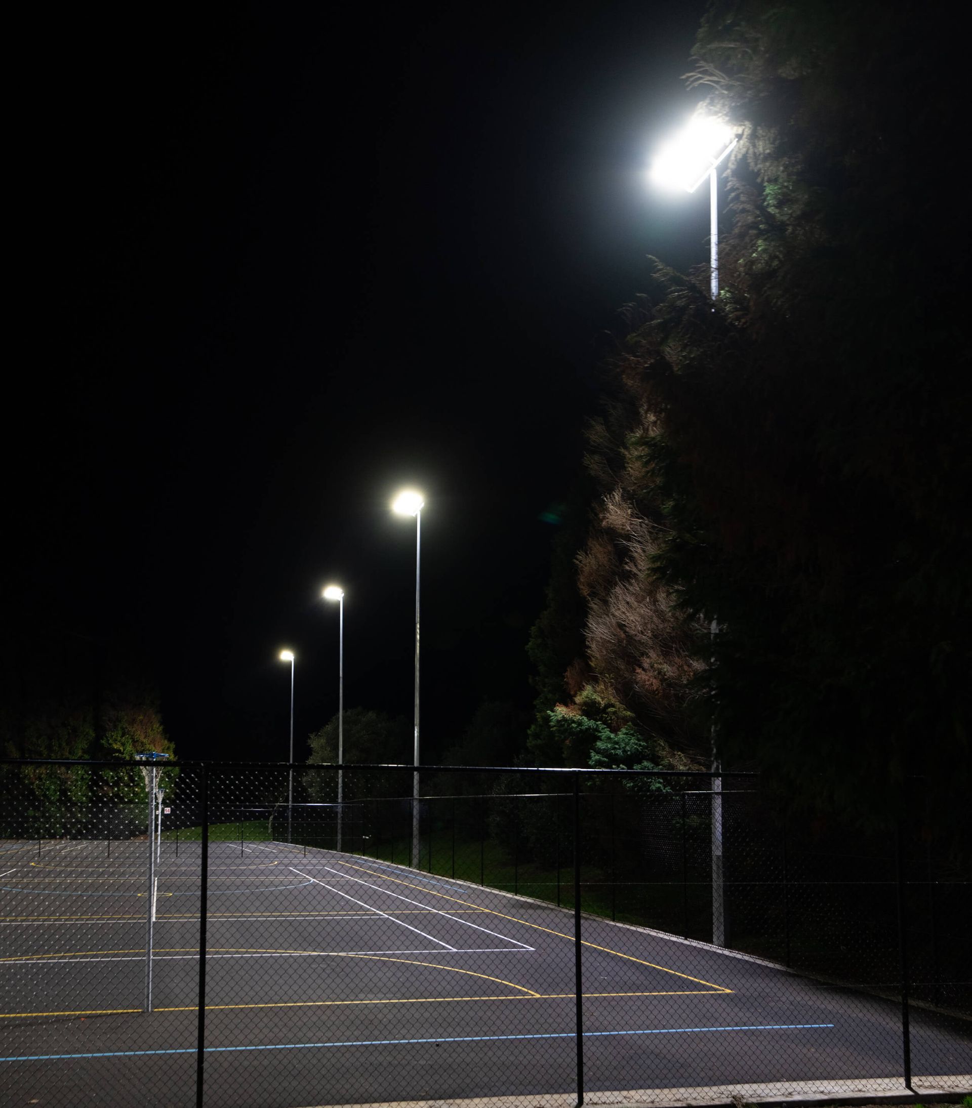 Nighttime view of a paved area with evenly spaced streetlights. A chain-link fence borders the area with dark, shadowy trees in the background.