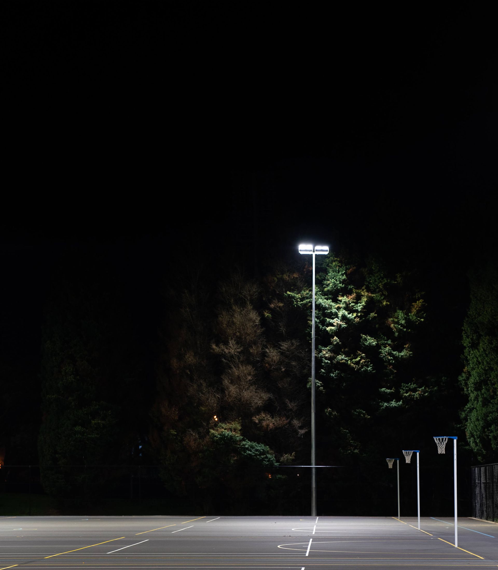Nighttime view of an empty basketball court under bright streetlights, with dark trees in the background.