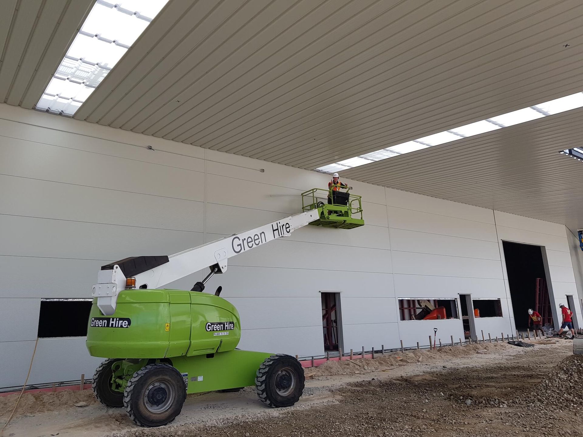 Green lift platform with worker near white building wall. Construction site setting with windows and exposed concrete.