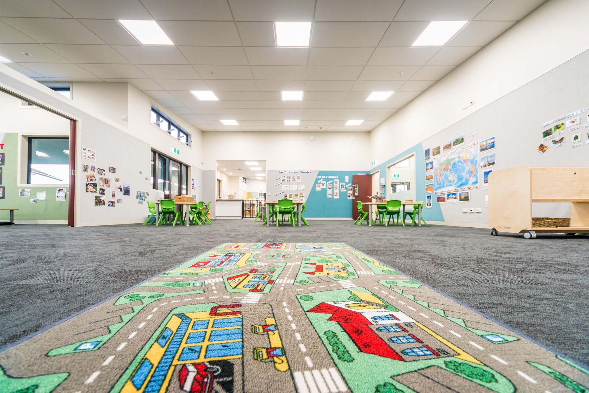A bright, empty classroom with a road rug in the foreground, tables, and artwork on the walls.