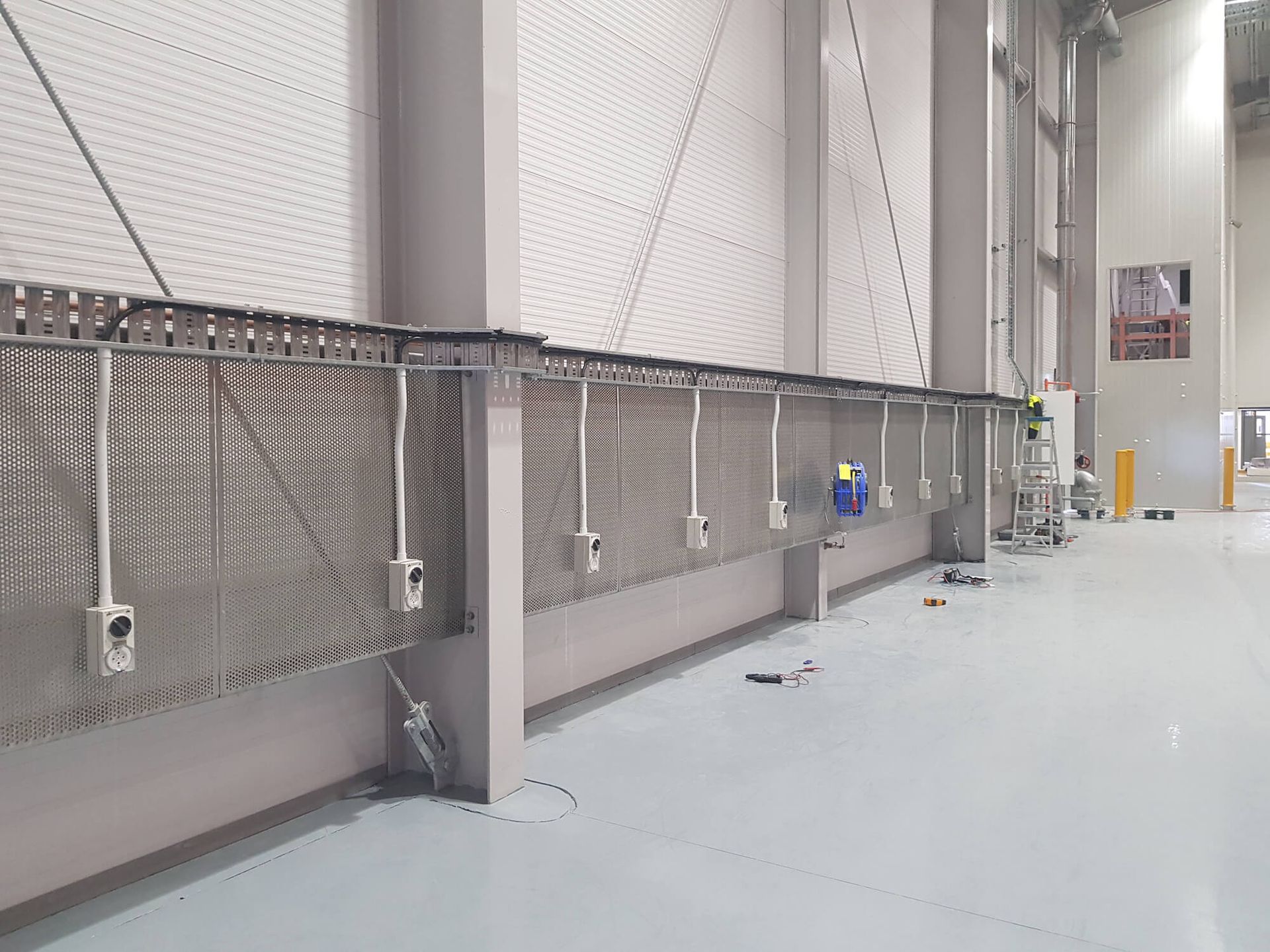 Interior view of a long, light-colored room with a metal mesh wall and electrical outlets lining the wall.