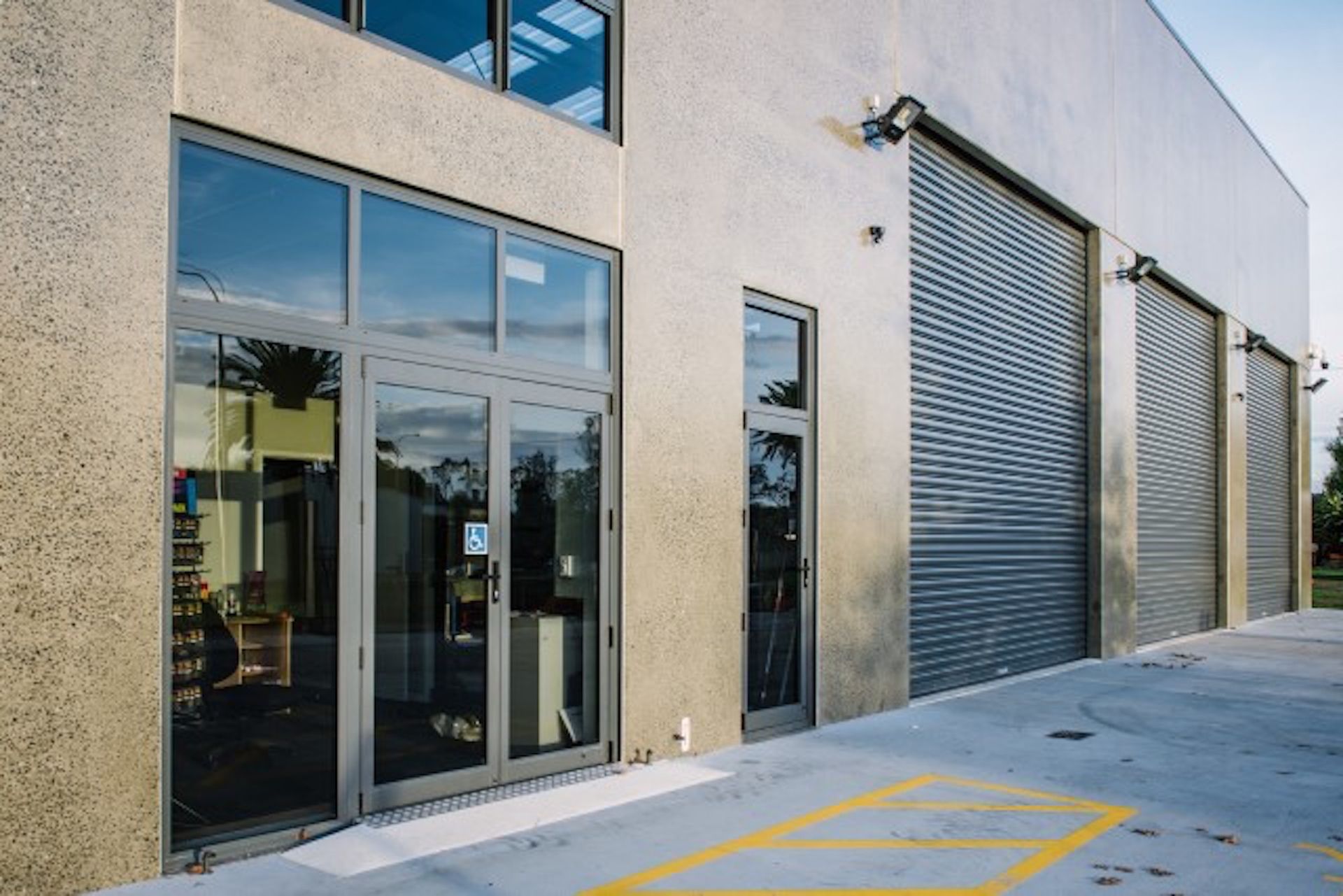 Exterior of a modern building with glass doors, a window, and three grey roller doors. Concrete walls, asphalt driveway, and a blue sky are visible.