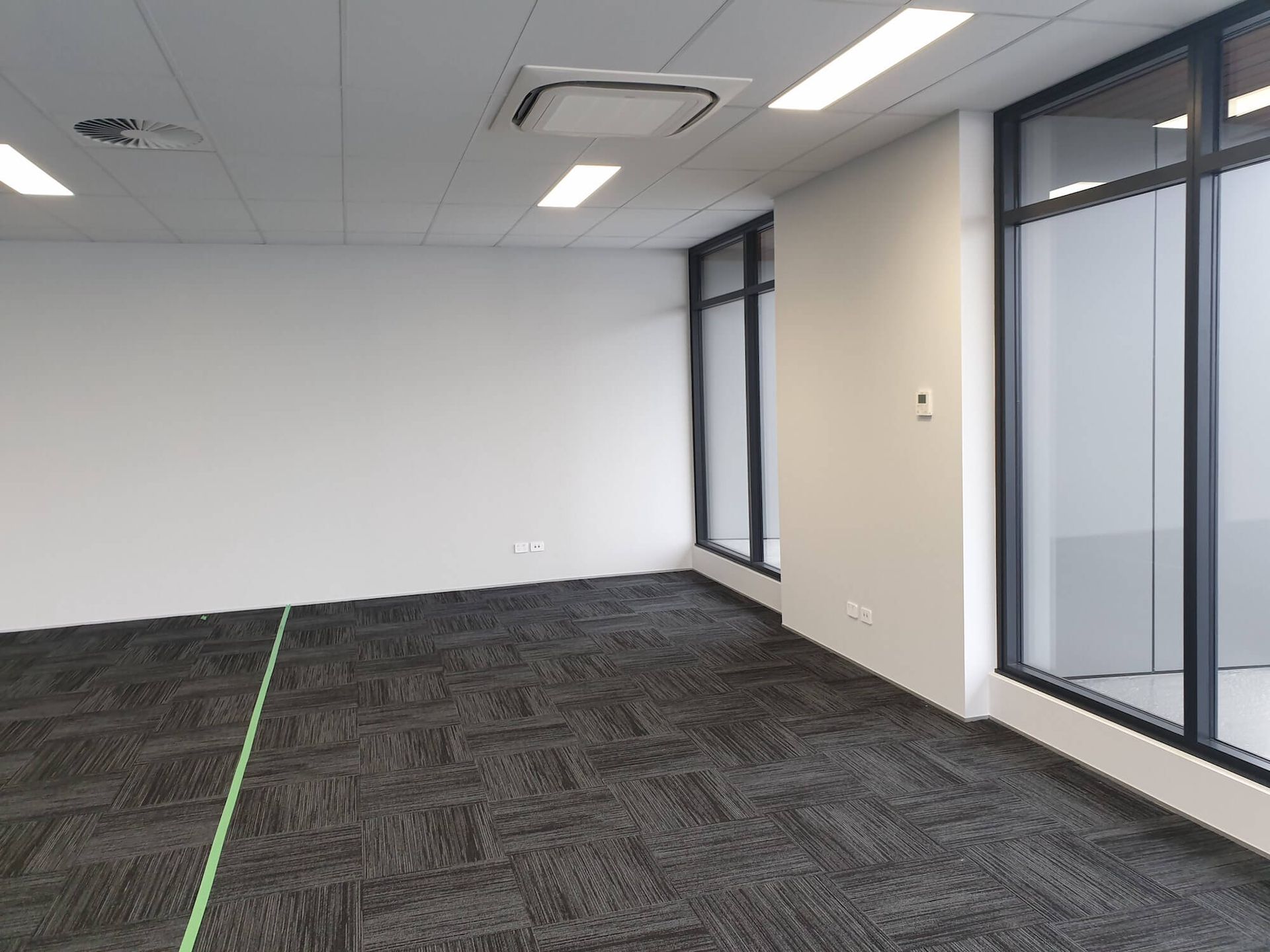 Empty office room with gray carpet, white walls, large windows, and fluorescent lights.