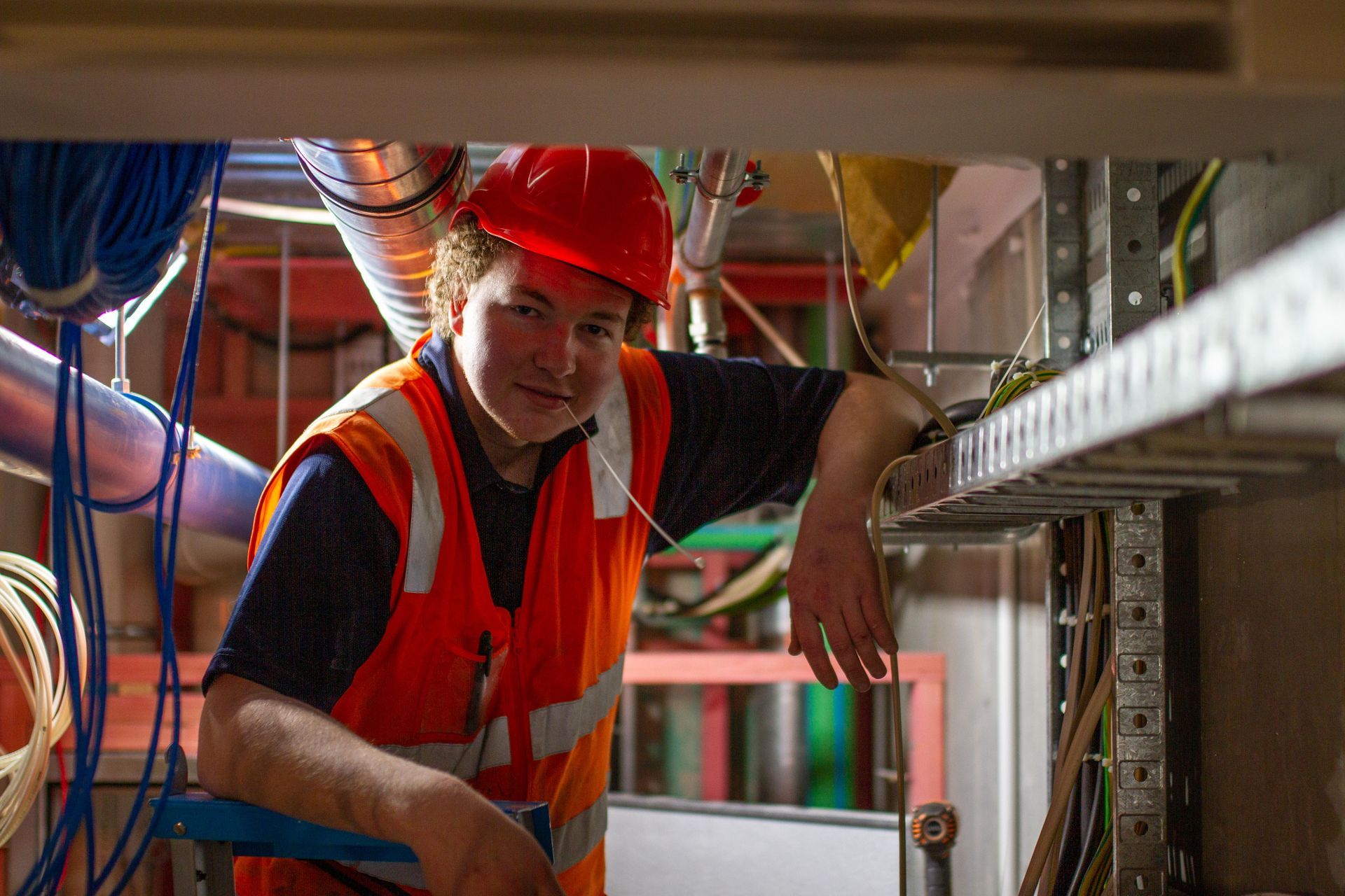 Construction worker in orange vest and hard hat smiles, leaning on a cable tray.