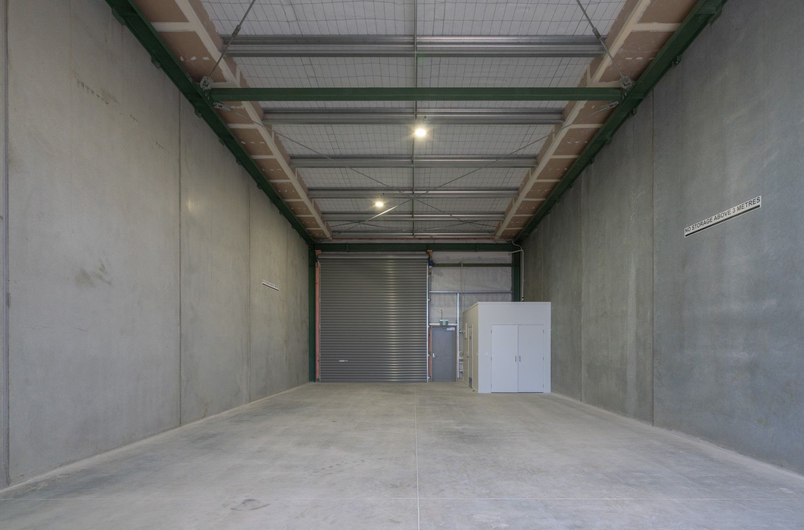 Empty storage unit with gray concrete walls and a metal roll-up door. Ceiling beams and lights are visible.