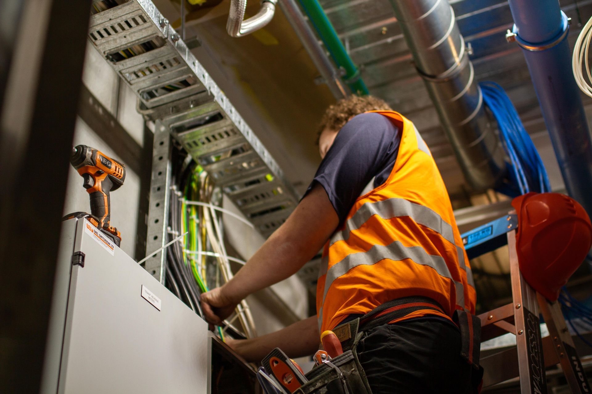 Electrician in orange safety vest works on wiring inside a building, with tools and cables visible.