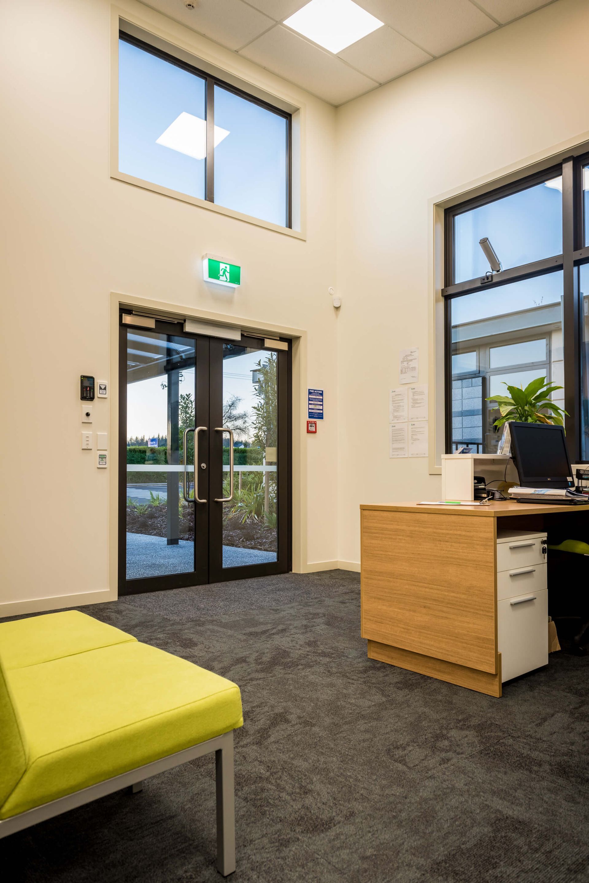 An office reception area with a green exit sign above glass doors, a desk with a computer, and a yellow bench. Grey carpet.