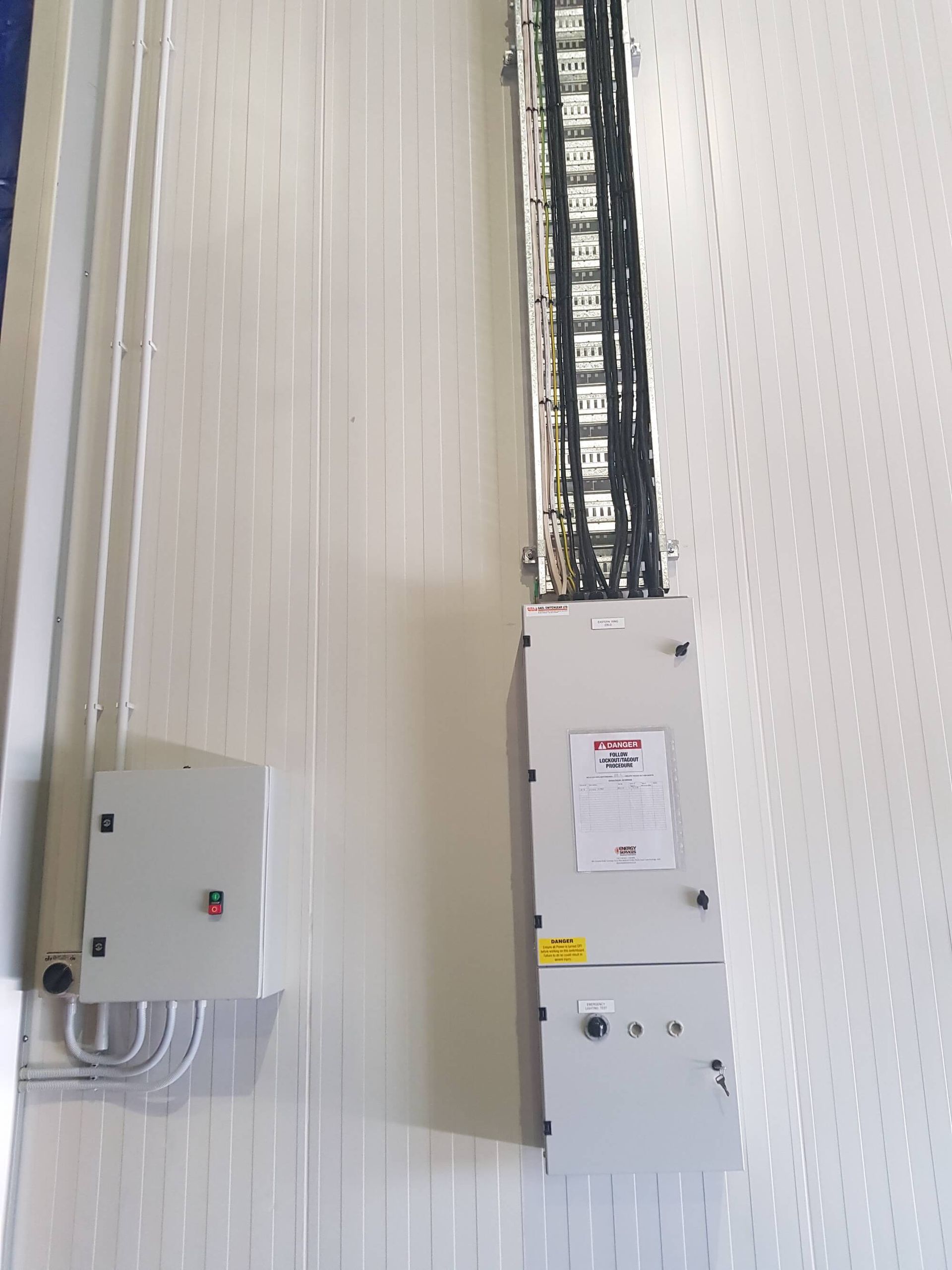Electrical boxes and wiring running vertically on a white wall, likely inside a building. A cable tray holds numerous wires.
