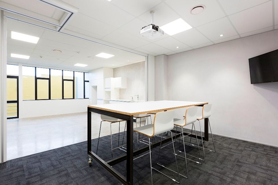 A modern meeting room with a large table and white chairs on a dark carpet. Sunlight streams in from a window.
