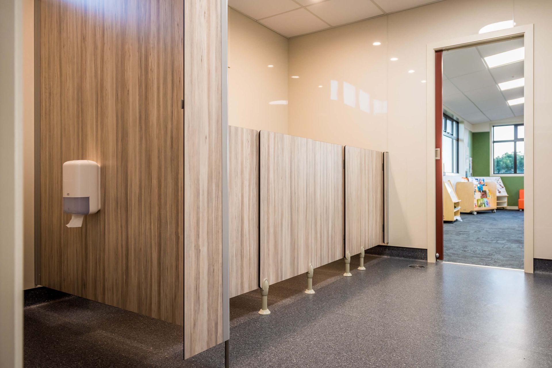 A public restroom with wood-grain stall dividers. A hand sanitizer dispenser is mounted on the wall. The door opens into another room.