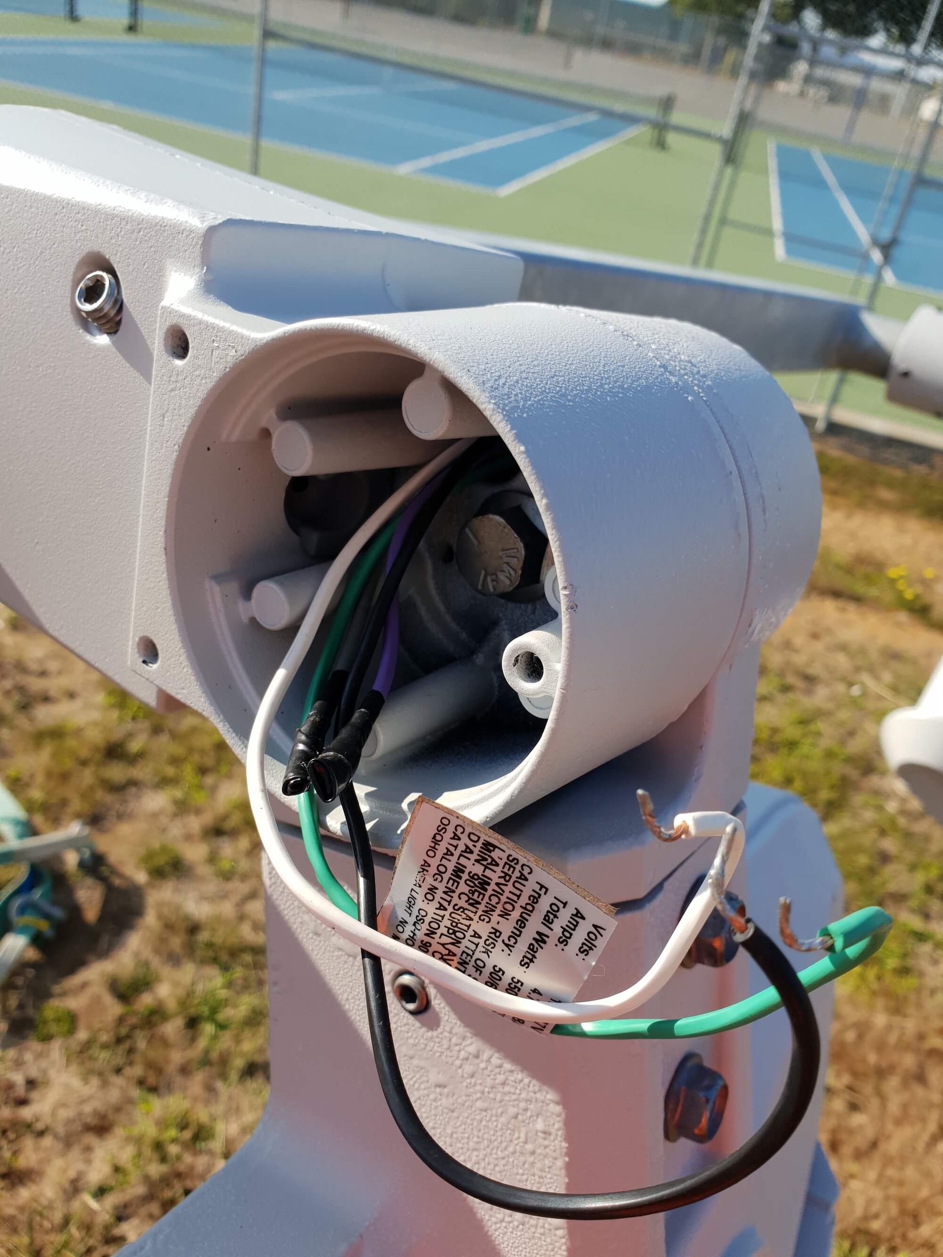 Open electrical conduit, showing wires and connections, near a tennis court. The wires are black, white, and green.