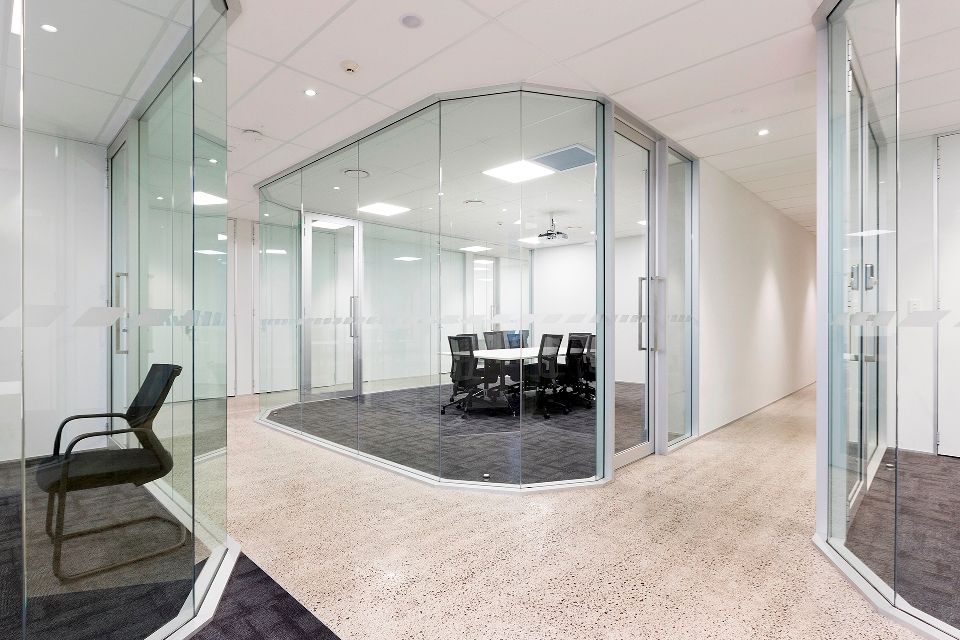 A modern office with glass-walled meeting rooms, a hallway, and black chairs. The rooms have a table, chairs, and a projector screen.