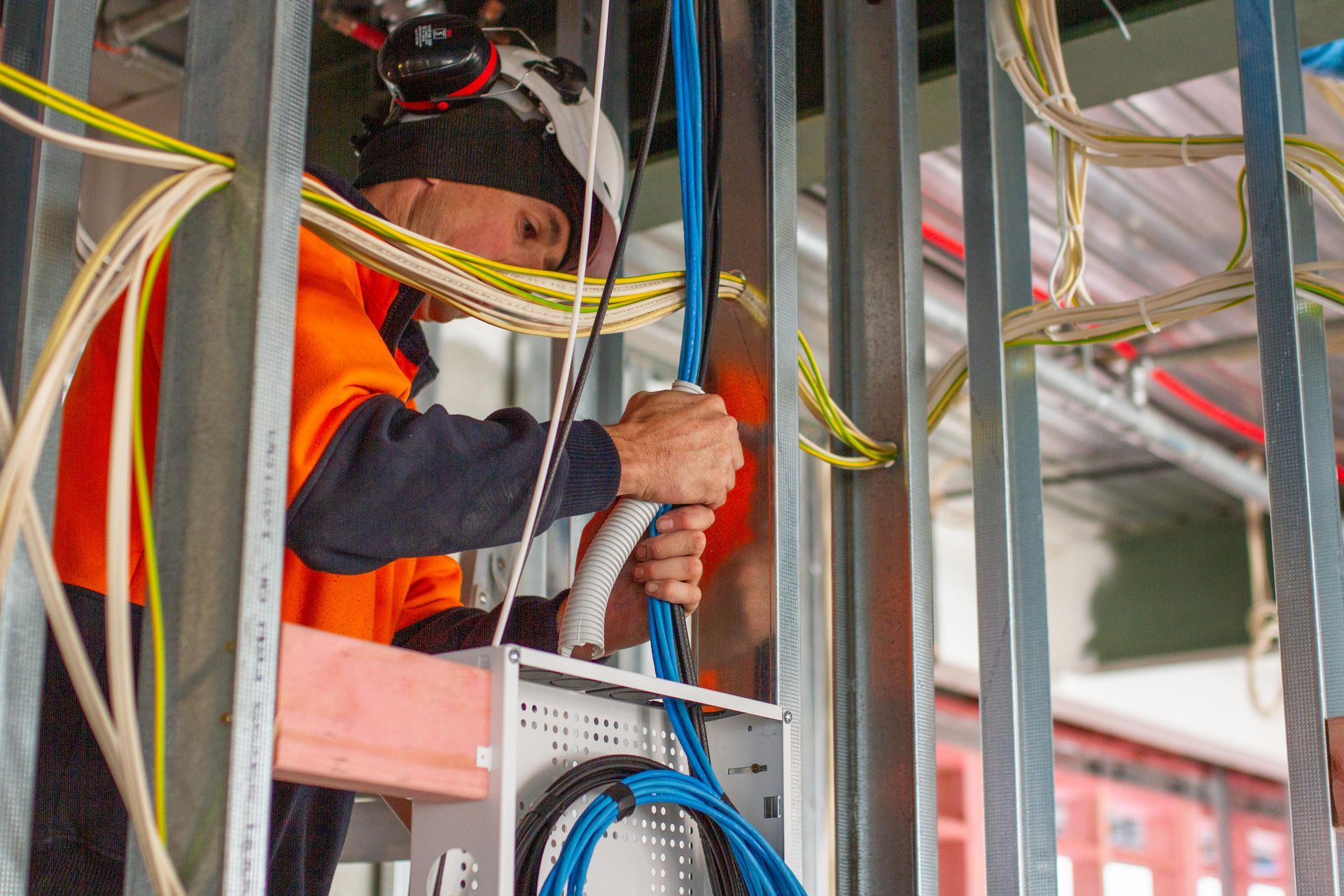 Electrician in orange safety vest working on wiring inside a metal frame wall.