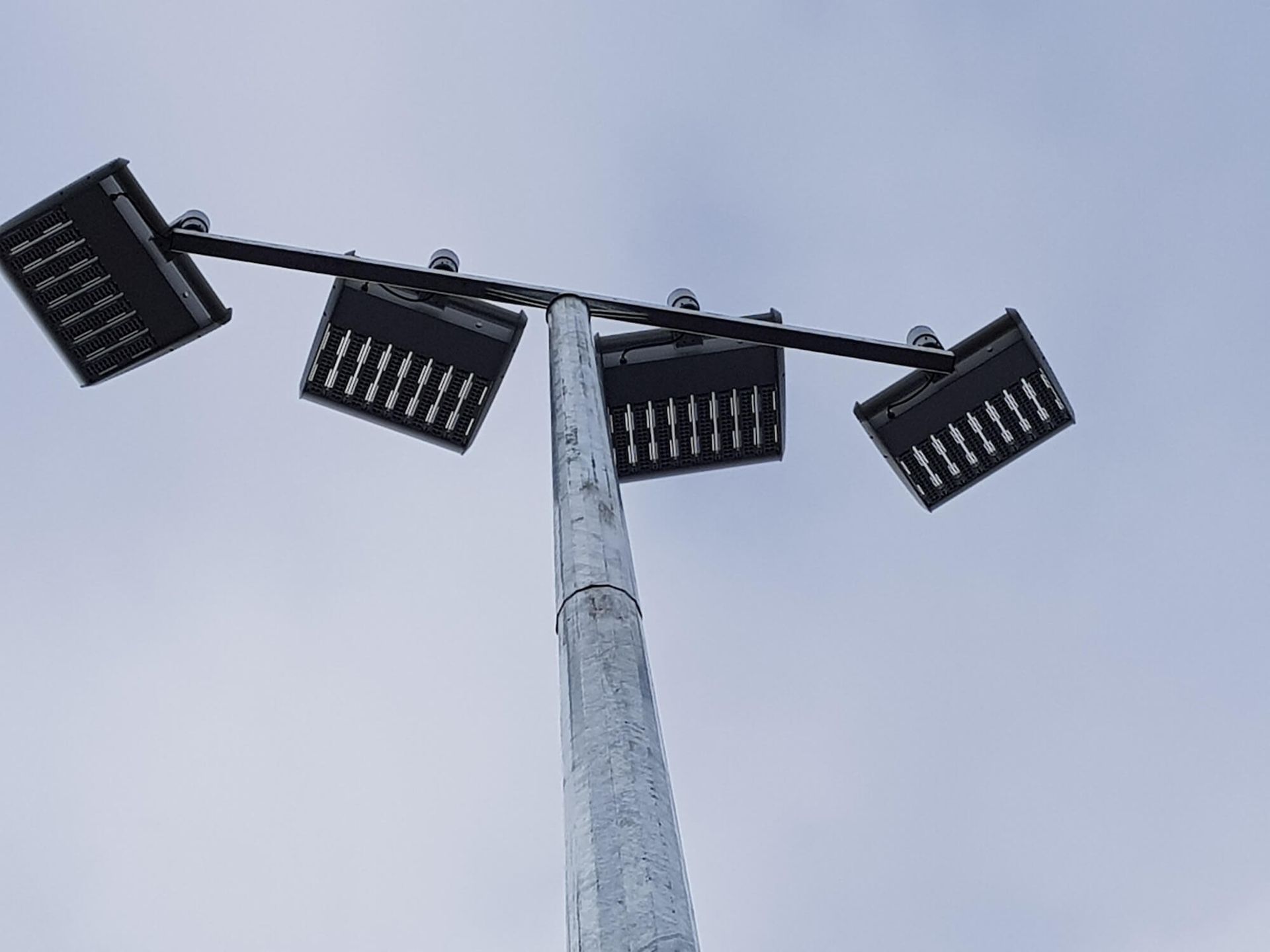 A tall metal pole with four square, black stadium lights angled towards the viewer against a cloudy sky.