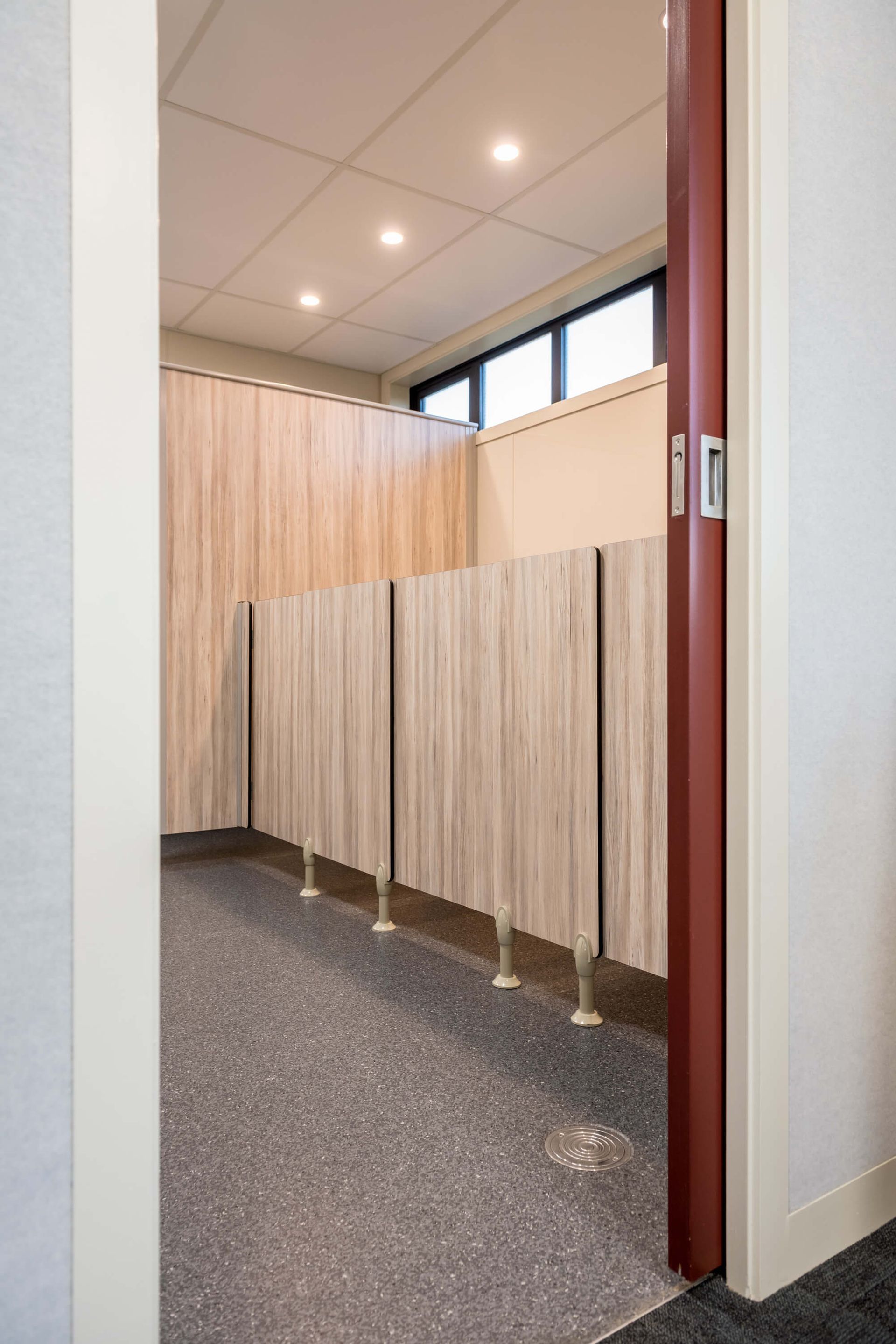 View from a doorway into a public restroom with light wood stalls, speckled floor, and a window.