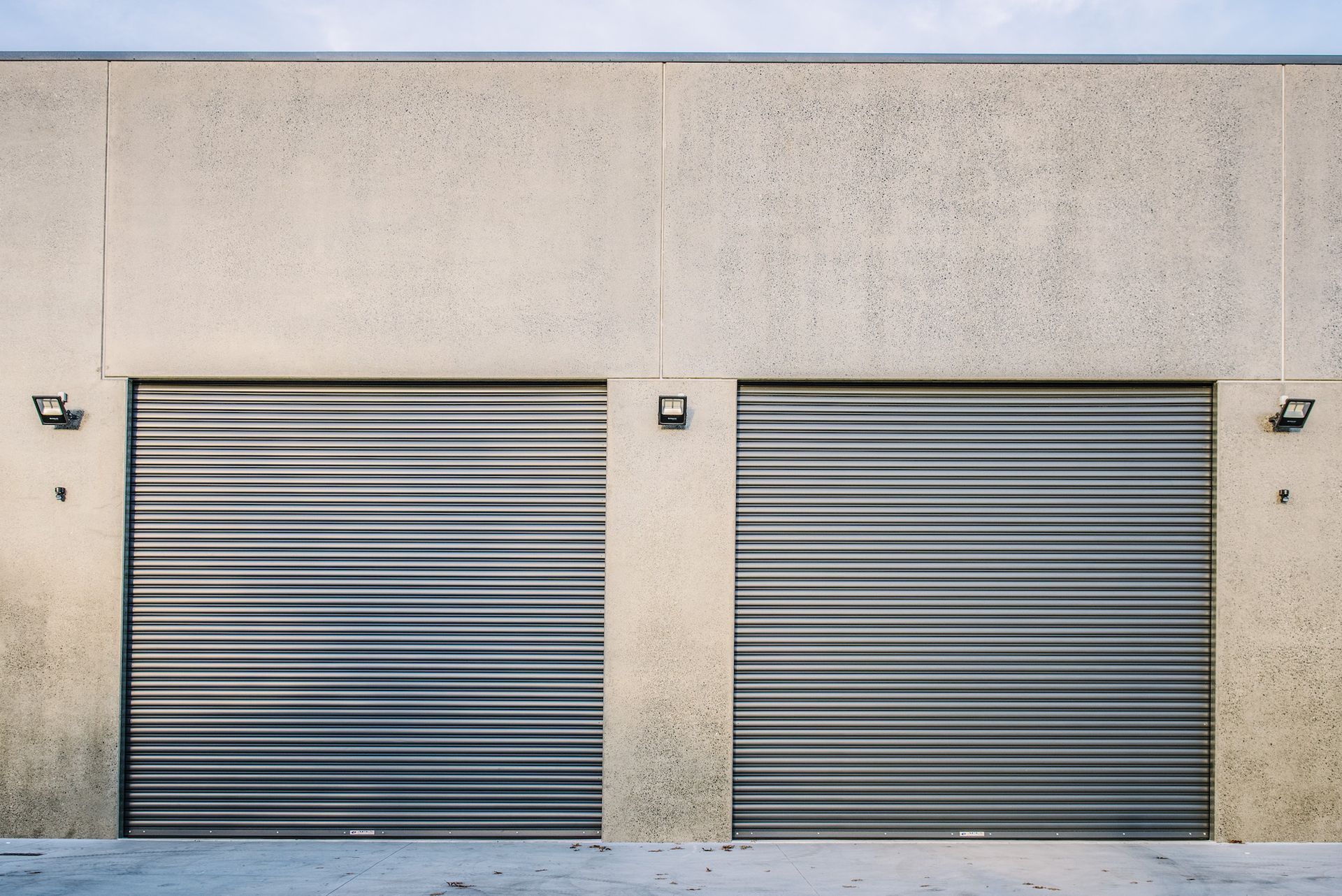 Two closed silver roll-up doors in a light-colored concrete wall. Two black lights are mounted above the doors.