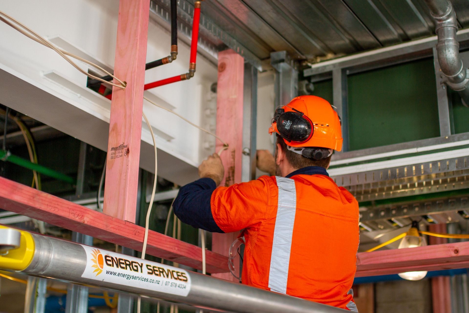 Construction worker in orange safety gear using tools to work on wood framing. Interior setting.