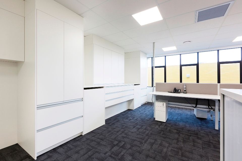 Modern office space with white cabinets and desks, dark carpet, and large windows.