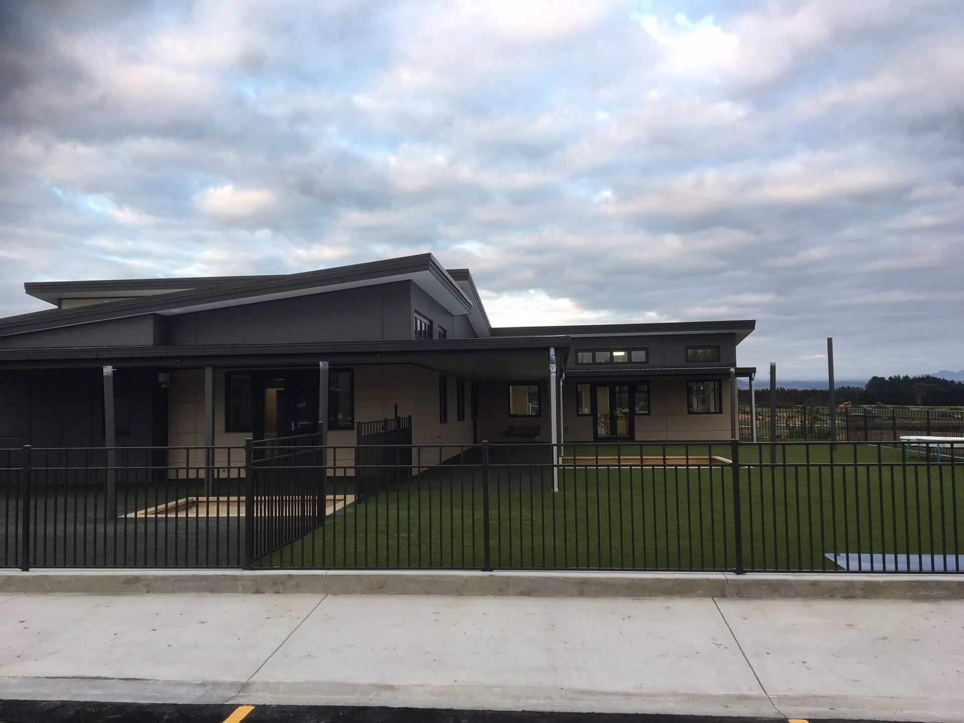 A modern, single-story building with a dark roof and a black fence in front, under a cloudy sky.