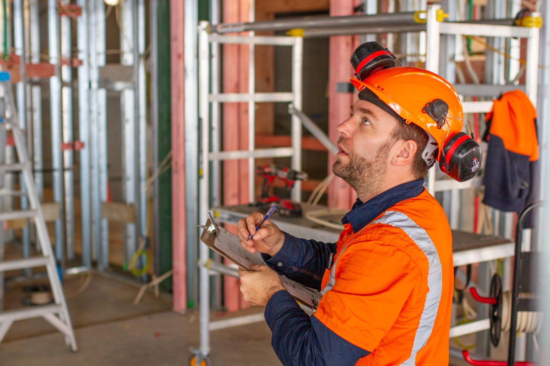 Construction worker in orange safety gear inspects a building site, writing on a clipboard. He wears a hard hat and ear protection.