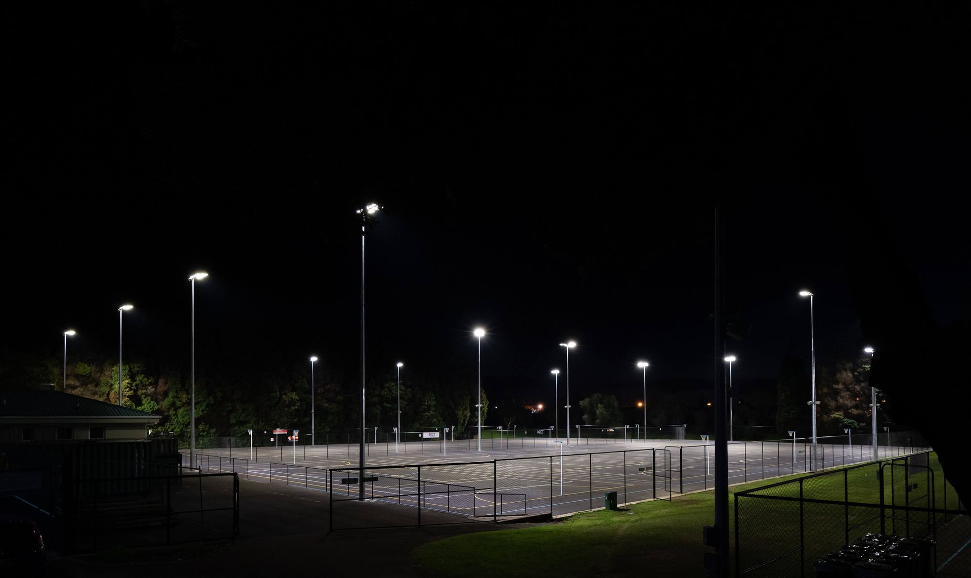 Nighttime view of an empty outdoor sports court illuminated by bright streetlights. Dark sky, green grass, and a fence are visible.