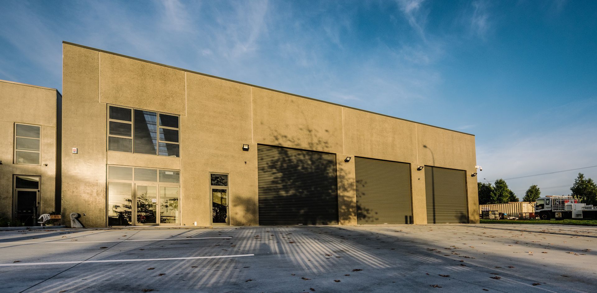 A light brown building with a sloped roof under a bright blue sky. Three garage doors are visible, and a concrete lot in front.