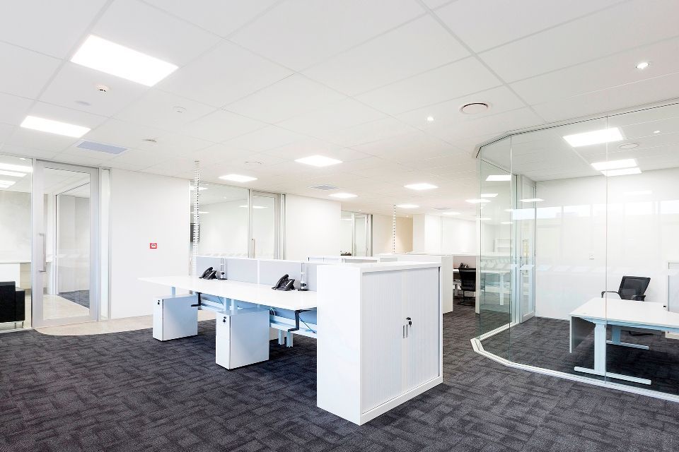 Bright office interior with white desks, cabinets, and walls. Dark gray carpet, glass partitions, and recessed lighting.