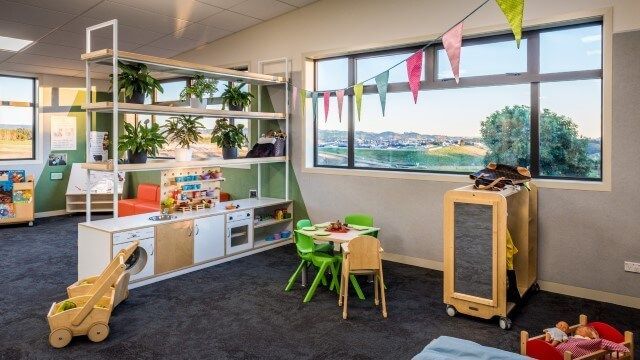 Childcare room with play kitchen, green chairs, and a large window overlooking a landscape with colorful bunting.