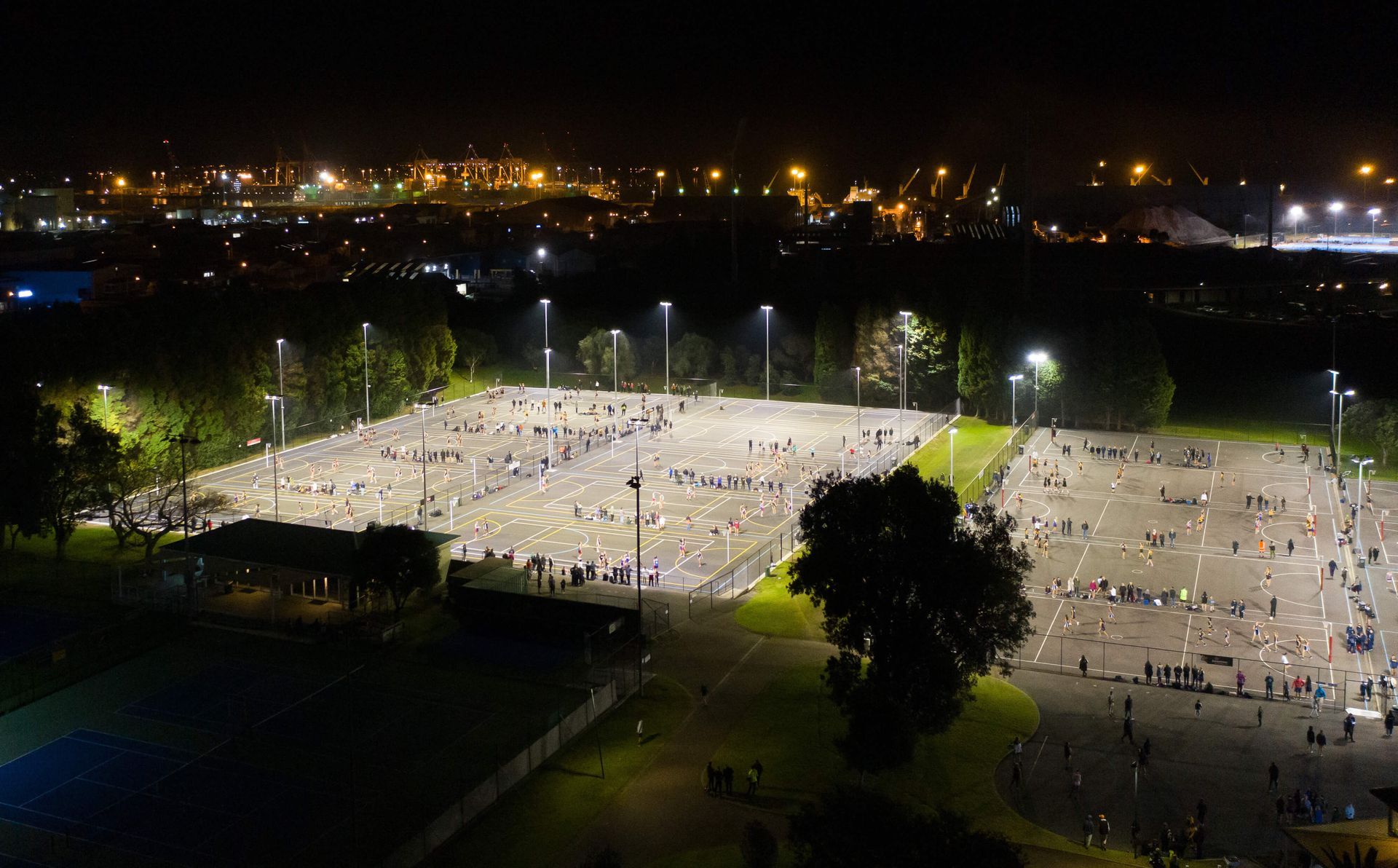 Nighttime aerial view of a large outdoor event. People are gathered on a lit, paved area, with surrounding trees and city lights in the background.