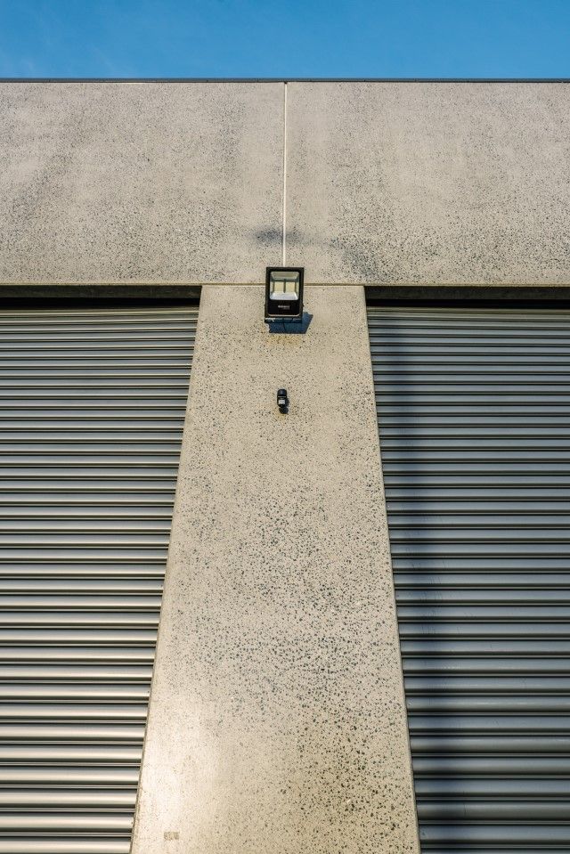 A gray building facade with two closed metal shutters. A spotlight and small camera are mounted above the shutters.