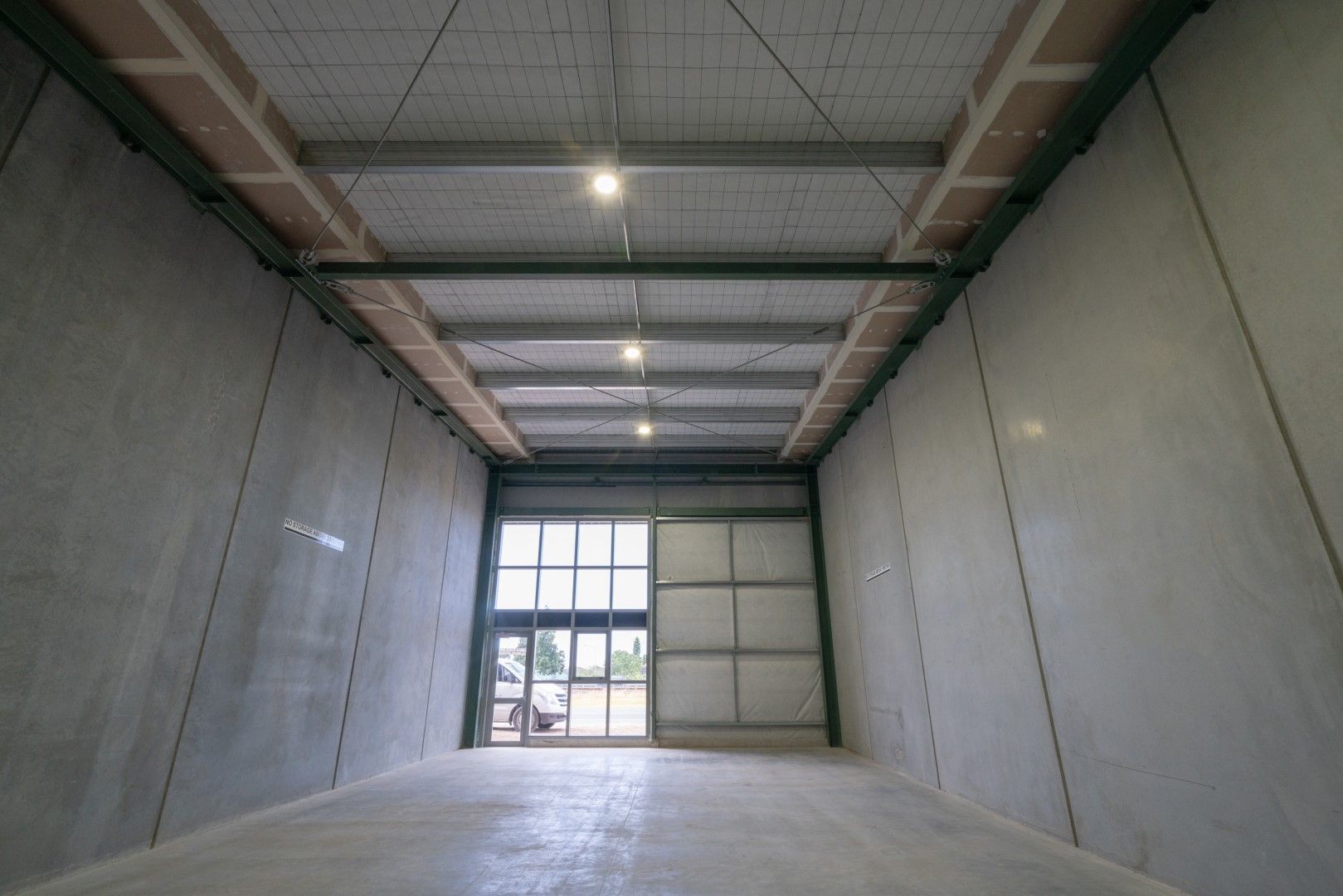 Interior of an empty storage unit with concrete walls, a closed garage door, and recessed lighting.