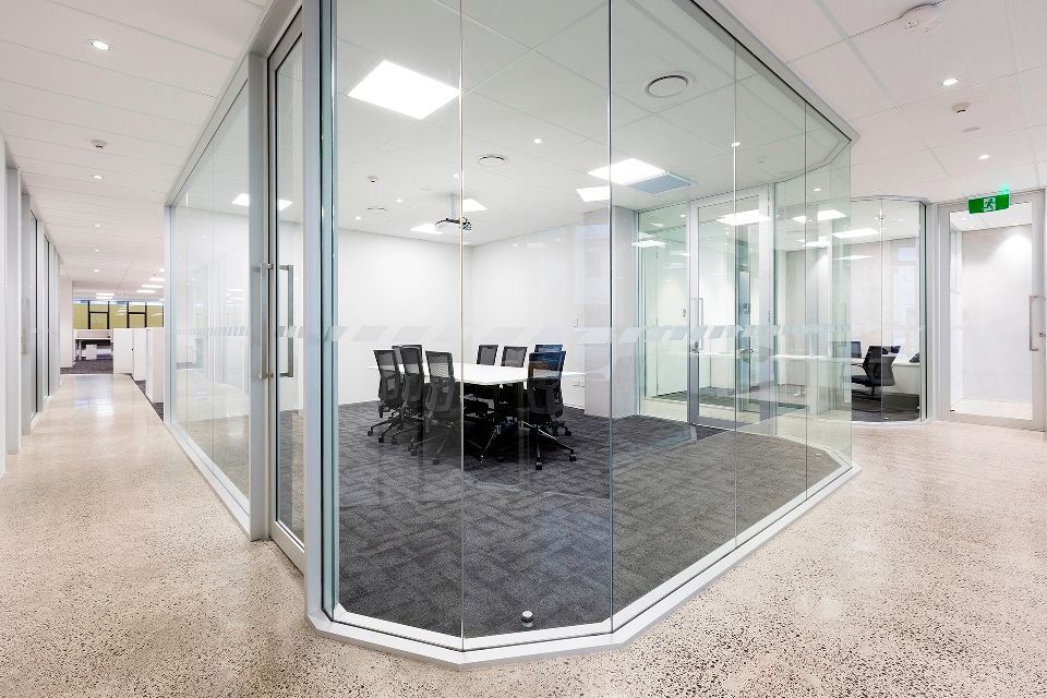 A modern office hallway with a glass-walled conference room. The room has a white table and black chairs; dark carpet and a bright white ceiling.