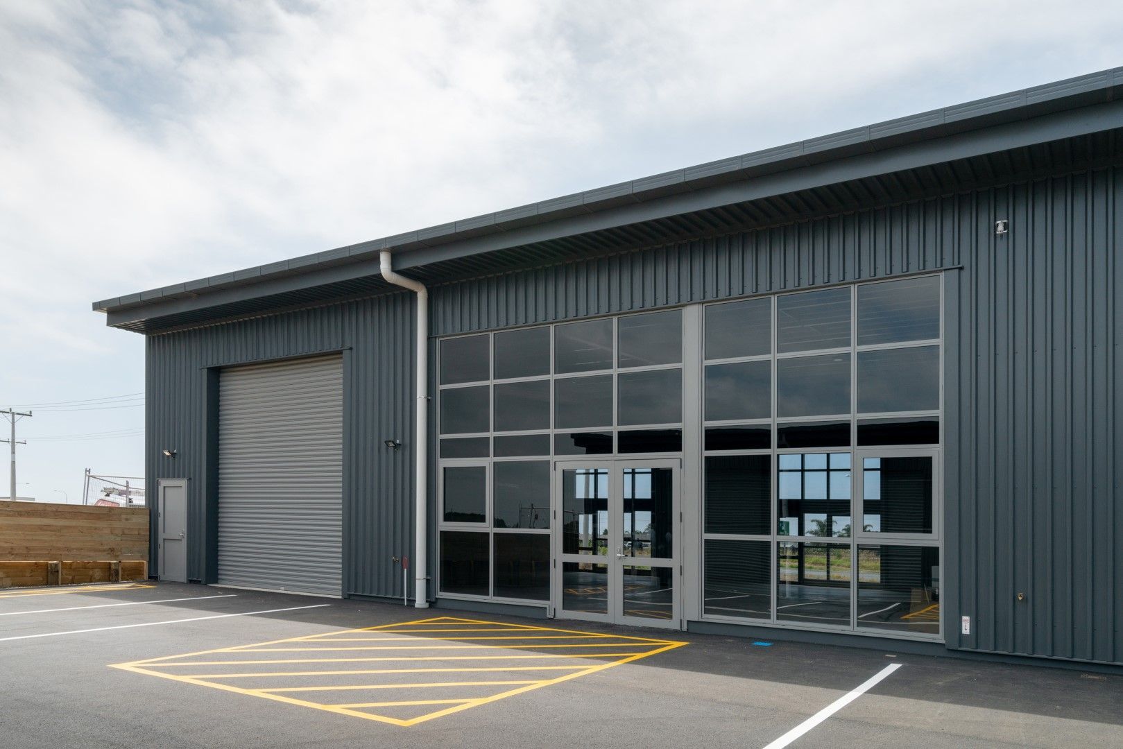 Gray commercial building with a large glass window and a garage door, set against a partly cloudy sky.