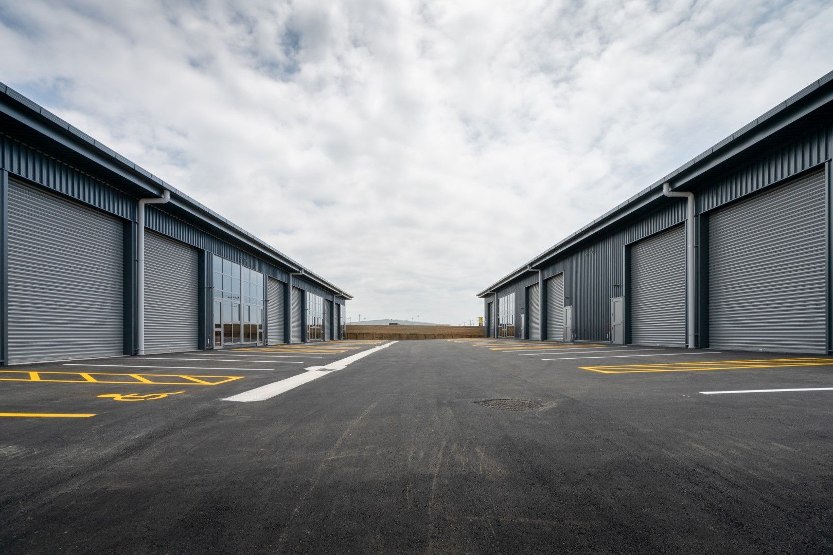 Two industrial buildings with roller doors face each other across an asphalt drive. The sky is overc