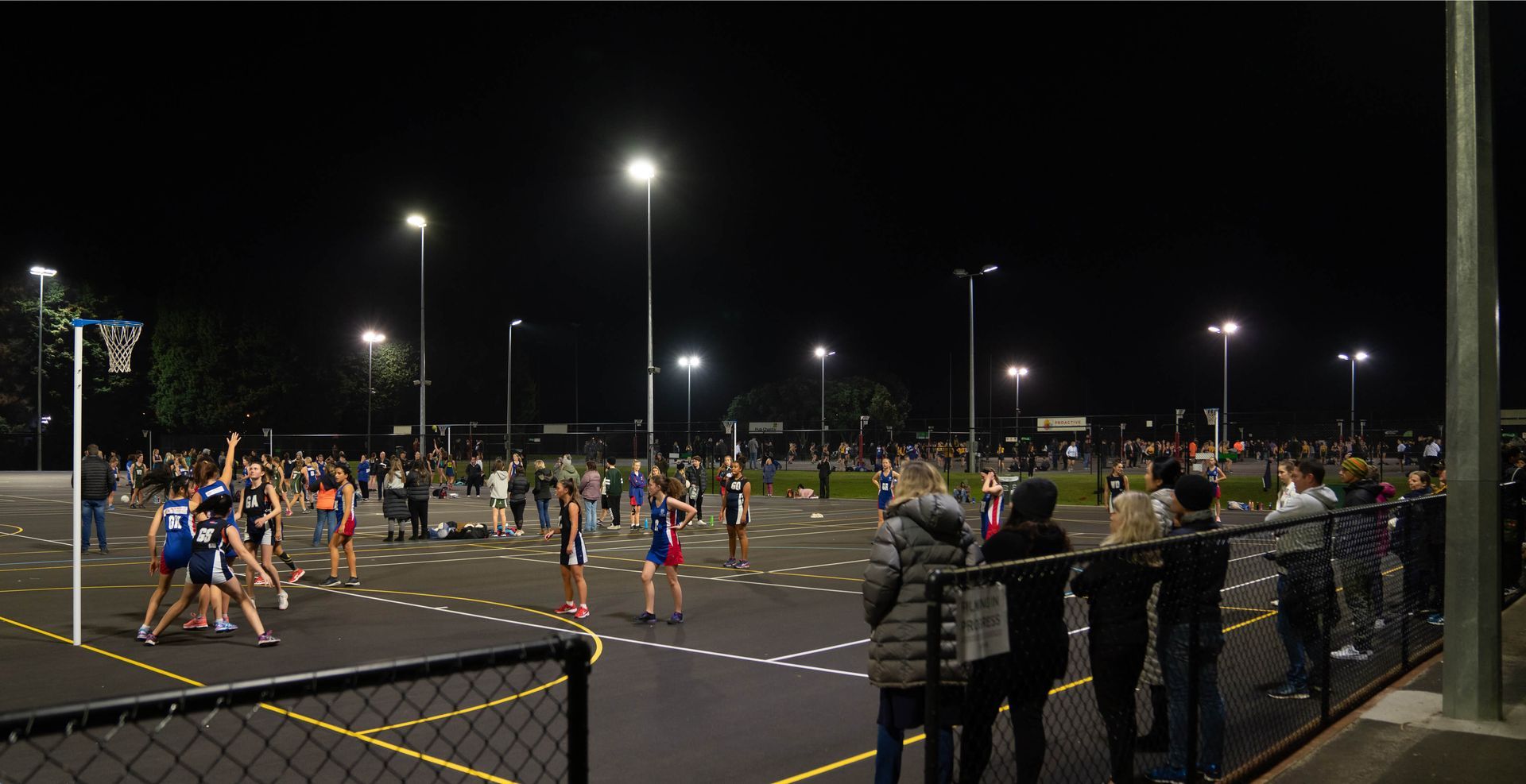 Netball game played at night under bright lights; players in uniforms on court, spectators watching behind a fence.