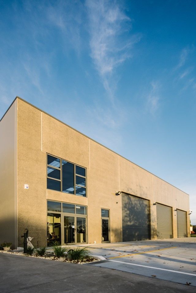A modern industrial building with tan brick walls, large windows, and three roll-up doors under a blue sky.