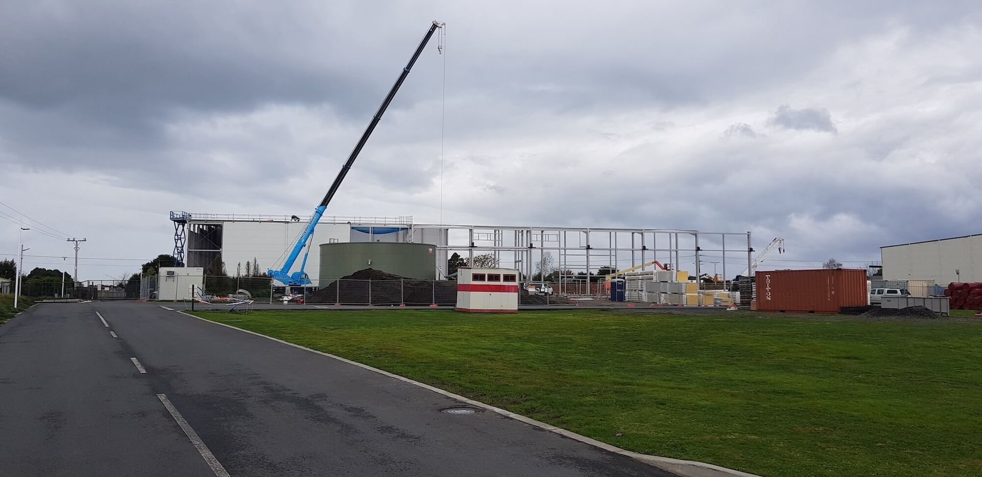 A construction site with a tall blue crane, metal framework, and containers under an overcast sky. A road and grassy area are in the foreground.
