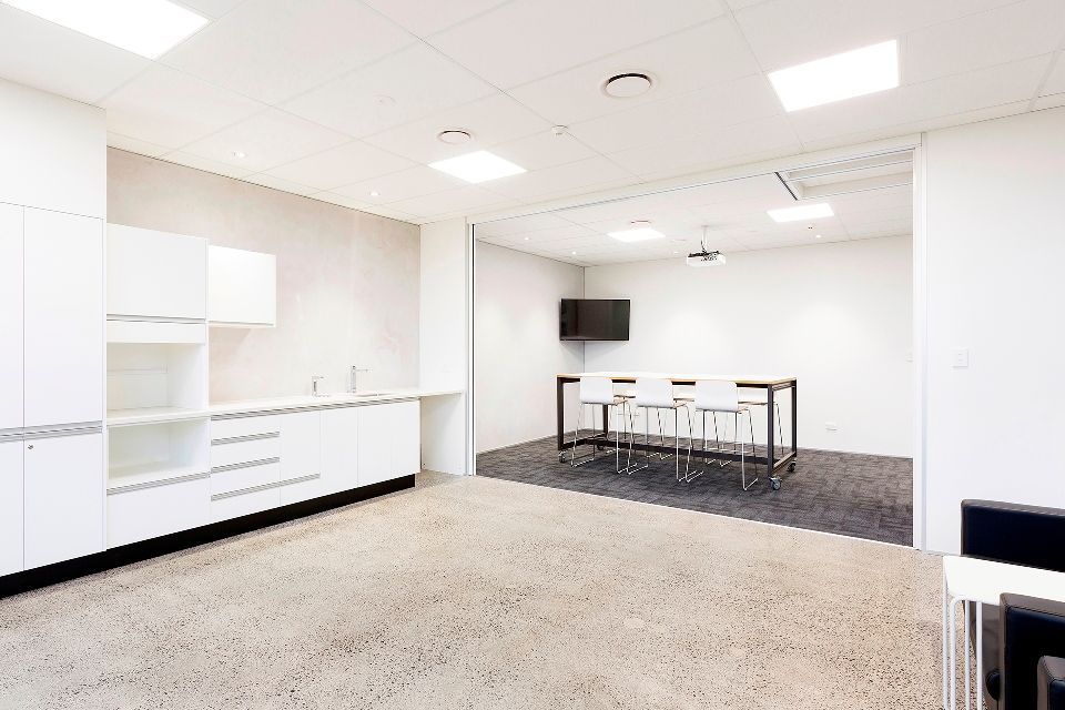 Bright, white office kitchen area with cabinets, countertops, and a table with chairs. A small TV is mounted on a wall.