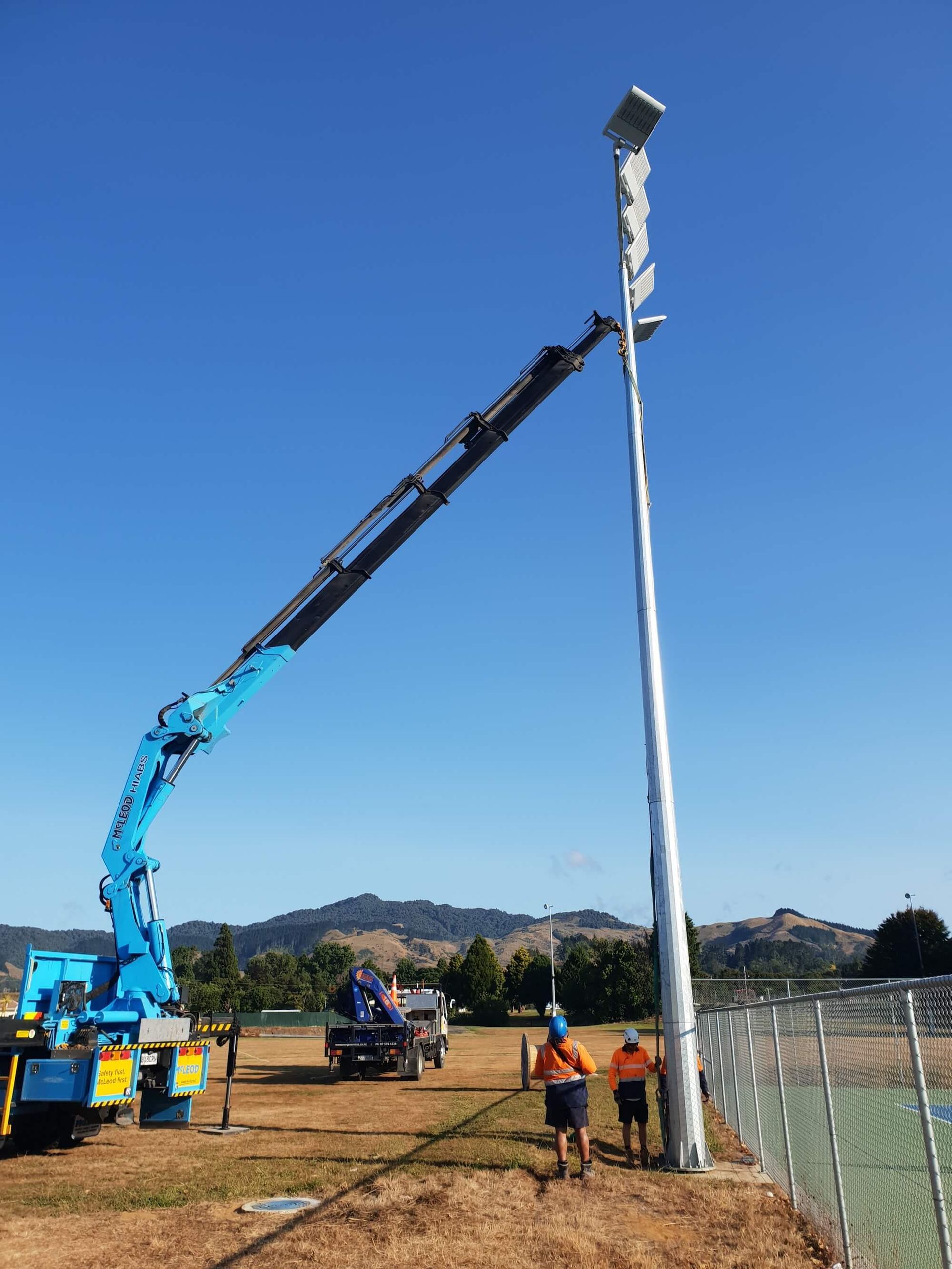 A blue crane is installing a tall light pole near a fence. Two workers are present on the ground under a bright blue sky.