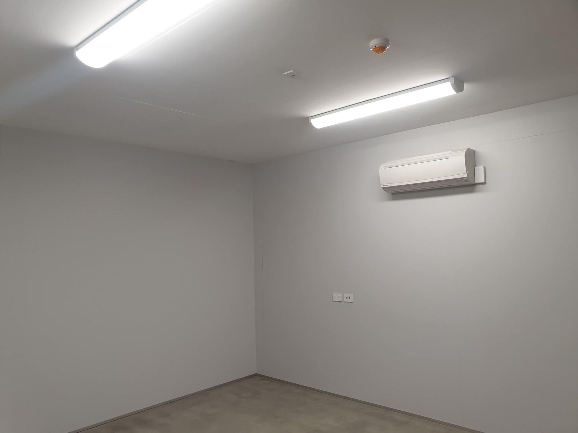 Empty, gray-walled room with concrete floor, two fluorescent lights, and an air conditioning unit.