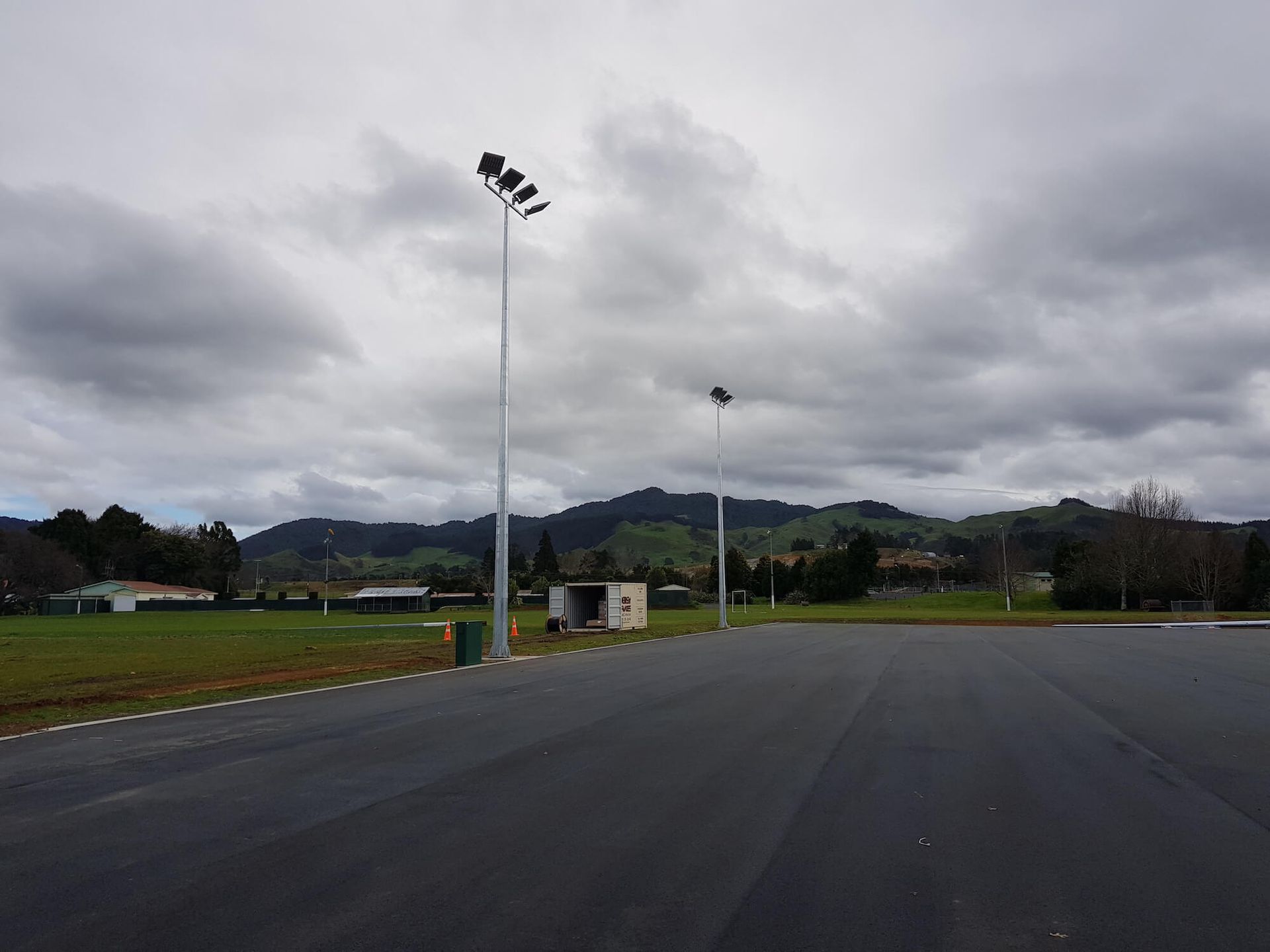 A parking lot with tall light poles against a backdrop of green hills and a cloudy sky.