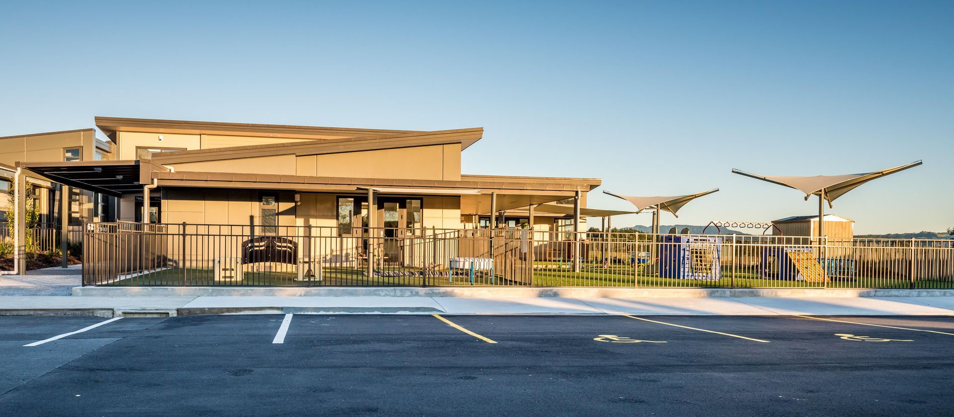A modern building with an outdoor shaded area, set against a clear blue sky. There is a parking lot in front of the building.